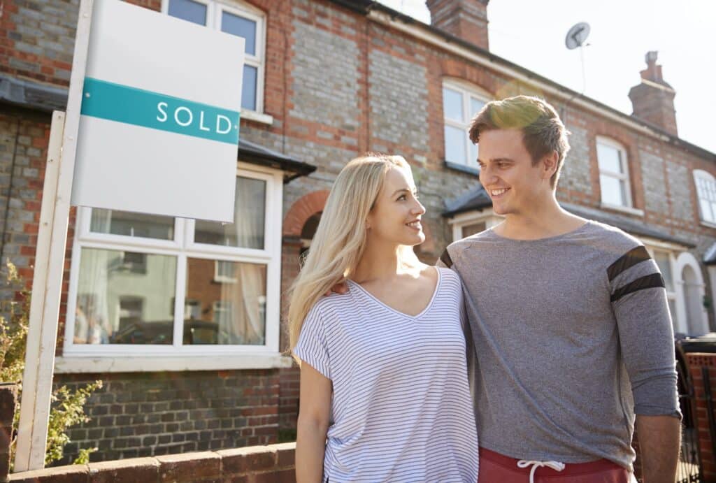 Excited Couple Standing Outside New Home With Sold Sign